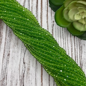 May include: A close-up of a strand of bright green beads. The beads are small, faceted, and strung together, creating a shimmering effect. The beads are arranged diagonally across a white and gray wooden surface. A green plant is in the background.