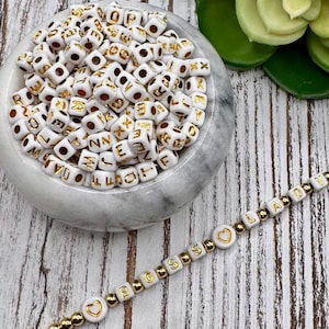 Puede incluir: Cuentas cúbicas blancas con letras doradas y una pulsera que deletrea "BOSS LADY". Las cuentas están en un cuenco de mármol y ensartadas en una cadena dorada. La pulsera incluye cuentas en forma de corazón.