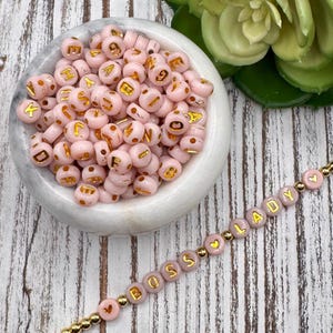 May include: Pink letter beads with gold lettering spelling "BOSS LADY" on a bracelet. A bowl of similar beads is in the background. The beads are round and have gold letters. The bracelet also has gold accent beads.