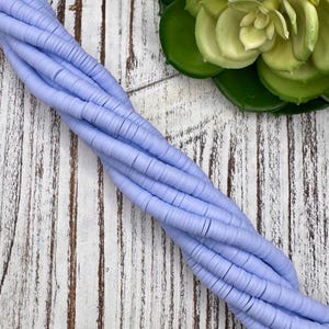 May include: A strand of light blue, flat polymer clay beads, ideal for jewelry making. The beads are arranged in a twisted pattern, showcasing their circular shape and uniform size. The background is a rustic, white-washed wooden surface.