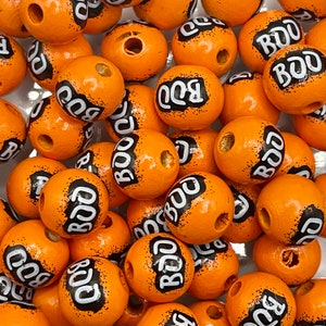 Puede incluir: Una colección de cuentas de madera naranja con letras negras que dicen "BOO" en cada cuenta. Las cuentas están dispuestas en un patrón aleatorio.