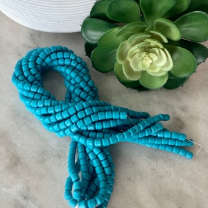 May include: A bundle of turquoise square beads, strung together, are displayed on a marble surface. The beads are a vibrant, solid color and appear to be made of glass or a similar material. A white ceramic vase and a green succulent plant are in the background.