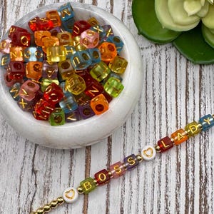 Könnte beinhalten: Eine Sammlung farbenfroher, durchscheinender Buchstabenperlen in einer weißen Schüssel, mit einem goldfarbenen Perlenarmband mit der Aufschrift "BOSS LADY". Die Perlen sind in verschiedenen Farben erhältlich, darunter Rot, Orange, Gelb, Grün und Blau.