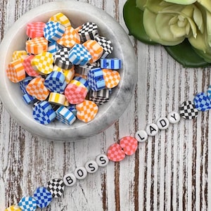 May include: A collection of colorful heart-shaped beads in a marble bowl, with a strand of beads spelling "BOSS LADY". The beads feature checkered patterns in various colors, including pink, blue, orange, yellow, and black.