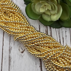 May include: A close-up of a large quantity of gold-colored round beads. The beads are arranged in a pile, creating a textured and shiny surface.