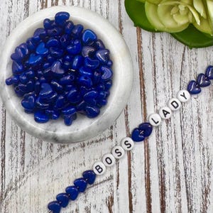Op de afbeelding: Een witte marmeren kom gevuld met glanzende, hartvormige, diepblauwe kralen. Een streng blauwe hartkralen spelt "BOSS LADY" met witte letterkralen. De kralen zijn gerangschikt op een wit, verweerd houten oppervlak.