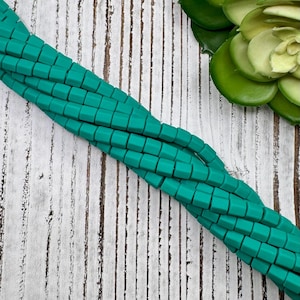 May include: A strand of teal green square-shaped beads. The beads are made of a matte material and have a slightly rounded edge. The beads are strung on a clear thread and are lying on a white wooden surface.