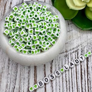 May include: A collection of white cube beads with green heart patterns in a white bowl. A strand of beads spells "BOSS LADY" with green heart accents. The beads are on a white, weathered wooden surface.