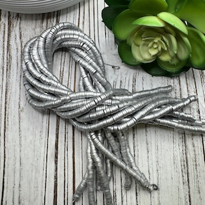May include: A strand of silver metal beads, each bead is a small circle. The beads are arranged in a loose coil on a white wooden surface.