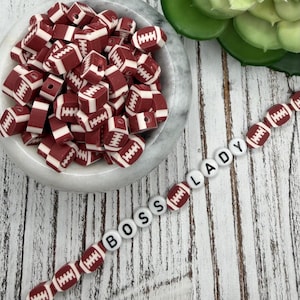 May include: A collection of maroon and white football-shaped beads in a marble bowl, with a strand of beads spelling "BOSS LADY". The beads are a mix of football shapes and round letter beads, perfect for crafting.
