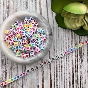 May include: White heart-shaped beads with rainbow colors inside, in a white bowl. A white bead bracelet with black letters spelling "BOSS LADY" is on a white wooden surface.