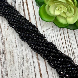 May include: A collection of black, faceted beads, possibly for jewellery, is shown against a light wood-effect backdrop. The beads have a shiny finish, reflecting light. A green plant is in the background.