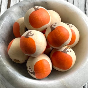 Puede incluir: Un tazón de cuentas de madera con un diseño de calabaza pintado en ellas. Las cuentas son de color marrón claro con calabazas naranjas pintadas en ellas.