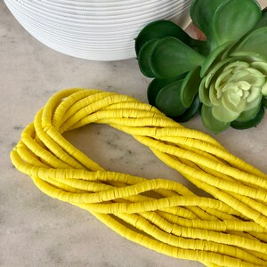 Puede incluir: Una hebra de cuentas de arcilla polimérica de color amarillo brillante. Las cuentas son redondas y planas, y están enhebradas juntas en una hebra larga y continua.