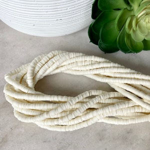 May include: A strand of off-white polymer clay beads. The beads are round and flat, and they are strung together in a long strand. The beads are arranged in a loose, flowing pattern.