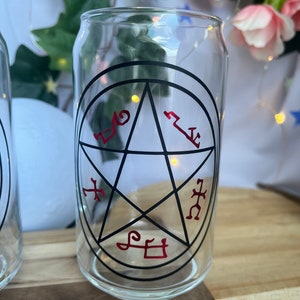 Pu&ograve; includere: Bicchiere trasparente a forma di lattina con un design a pentagramma nero e simboli rossi. Il bicchiere &egrave; vuoto e poggia su una superficie di legno, con vegetazione e fiori sullo sfondo.