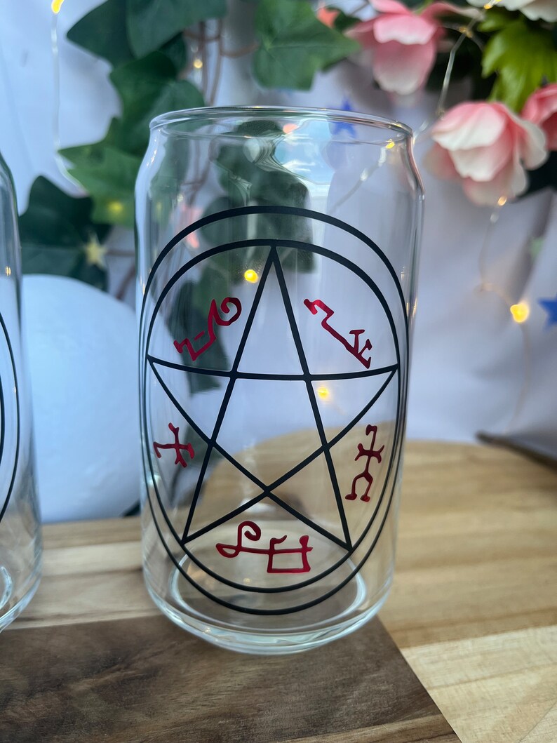 Pu&ograve; includere: Bicchiere trasparente a forma di lattina con un design a pentagramma nero e simboli rossi. Il bicchiere &egrave; vuoto e poggia su una superficie di legno, con vegetazione e fiori sullo sfondo.