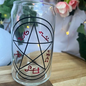 Pu&ograve; includere: Un bicchiere trasparente a forma di lattina con un design a pentagramma nero e simboli rossi. Il bicchiere &egrave; appoggiato su una superficie di legno, con uno sfondo sfocato di fiori e verde.