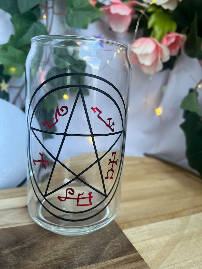 Pu&ograve; includere: Un bicchiere trasparente a forma di lattina con un design a pentagramma nero e simboli rossi. Il bicchiere &egrave; appoggiato su una superficie di legno, con uno sfondo sfocato di fiori e verde.