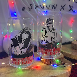Könnte beinhalten: Zwei transparente Trinkgläser in Dosenform mit schwarz-weißen Charakterillustrationen. Der Text "THE BOY WITH THE BATS" ist rot auf der Unterseite jedes Glases aufgedruckt. Die Gläser stehen auf einer Holzoberfläche mit Lichterketten.