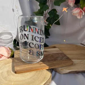 Può includere: Un bicchiere trasparente a forma di lattina con la scritta "RUNNING ON ICED COFFEE & SMILES" in nero. Il bicchiere è appoggiato su un piccolo tagliere di legno. Lo sfondo comprende vegetazione e luci fatate.