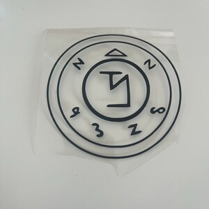 Pu&ograve; includere: Adesivo circolare nero con un design a sigillo. L'adesivo presenta un simbolo centrale all'interno di un cerchio, circondato da altri simboli e numeri. L'adesivo &egrave; su un supporto trasparente, pronto per l'applicazione.