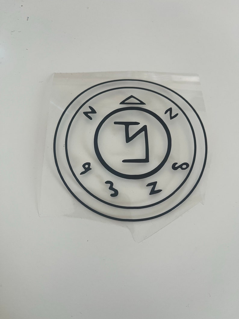 Pu&ograve; includere: Adesivo circolare nero con un design a sigillo. L'adesivo presenta un simbolo centrale all'interno di un cerchio, circondato da altri simboli e numeri. L'adesivo &egrave; su un supporto trasparente, pronto per l'applicazione.