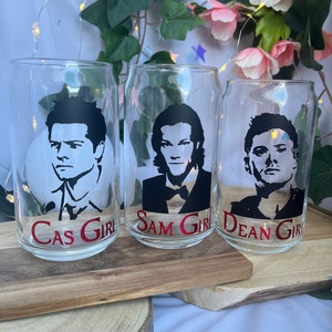 Könnte beinhalten: Drei transparente Gläser mit schwarzen Silhouetten männlicher Figuren. Jedes Glas hat einen roten Text am Boden: "Cas Girl", "Sam Girl" und "Dean Girl". Die Gläser stehen auf einem Holztablett.
