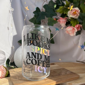 Könnte beinhalten: Ein klares, becherförmiges Trinkglas mit dem Text "I LIKE MY BOOKS SPICY AND MY COFFEE ICY" in schwarzer und holografischer Schrift. Das Glas zeigt eine Grafik mit einem Bücherstapel und wird auf einem Holzschneidebrett präsentiert.
