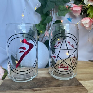 Pu&ograve; includere: Due bicchieri trasparenti a forma di lattina con disegni grafici neri e rossi. Un bicchiere presenta un simbolo rosso all'interno di un cerchio nero, l'altro un pentagramma nero con simboli rossi. I bicchieri sono su un vassoio di legno.