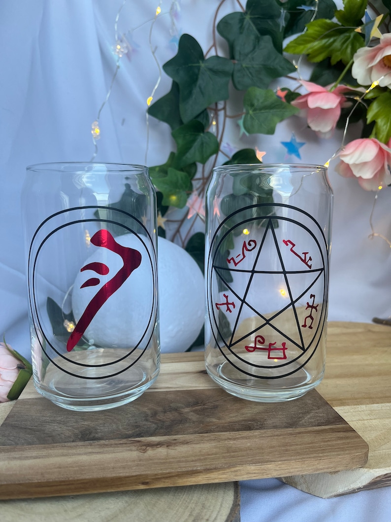 Pu&ograve; includere: Due bicchieri trasparenti a forma di lattina con disegni grafici neri e rossi. Un bicchiere presenta un simbolo rosso all'interno di un cerchio nero, l'altro un pentagramma nero con simboli rossi. I bicchieri sono su un vassoio di legno.