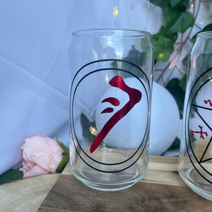 Pu&ograve; includere: Bicchiere trasparente a forma di lattina con un simbolo rosso all'interno di un cerchio nero. Il bicchiere &egrave; su una superficie di legno. Lo sfondo include tessuto bianco, luci di fata e vegetazione.