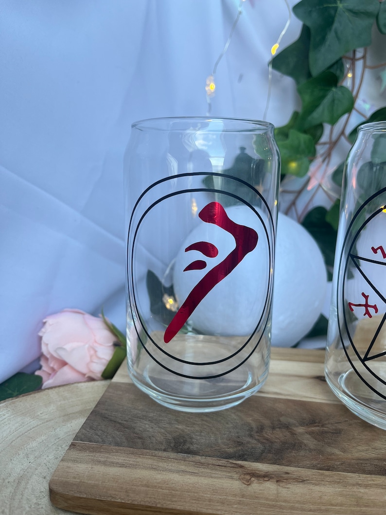 Pu&ograve; includere: Bicchiere trasparente a forma di lattina con un simbolo rosso all'interno di un cerchio nero. Il bicchiere &egrave; su una superficie di legno. Lo sfondo include tessuto bianco, luci di fata e vegetazione.