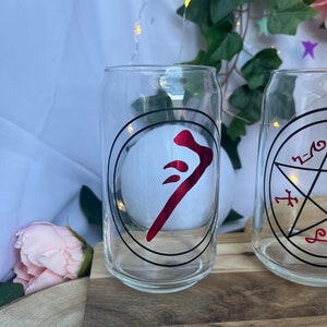 Pu&ograve; includere: Due bicchieri trasparenti a forma di lattina con disegni circolari neri e simboli rossi. Un bicchiere presenta un simbolo rosso all'interno di un cerchio nero, l'altro un disegno a pentagramma nero. I bicchieri sono su una superficie di legno.