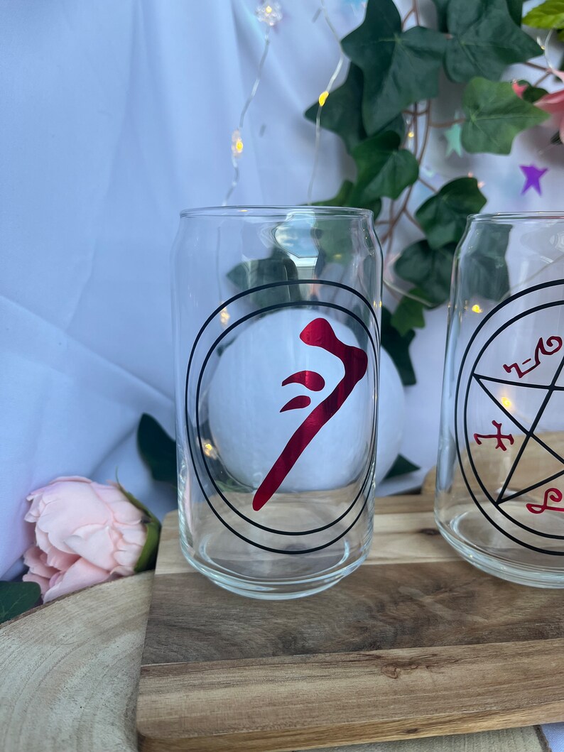 Pu&ograve; includere: Due bicchieri trasparenti a forma di lattina con disegni circolari neri e simboli rossi. Un bicchiere presenta un simbolo rosso all'interno di un cerchio nero, l'altro un disegno a pentagramma nero. I bicchieri sono su una superficie di legno.