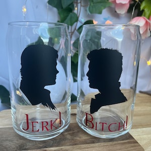 May include: Two clear glass can-shaped drinking glasses. Each glass features a black silhouette of a person's profile. The word "Jerk!" is printed in red on one glass, and "Bitch!" is printed in red on the other. The glasses are on a wooden surface.