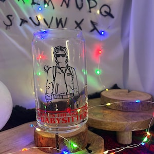 Könnte beinhalten: Ein klares, becherförmiges Trinkglas mit einer schwarzen Grafik eines Charakters mit Sonnenbrille und Jacke. Der Text "ALWAYS THE GOOD DAD BABYSITTER" ist rot aufgedruckt. Das Glas steht auf einem Holzuntersetzer.