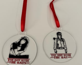 Puede incluir: Dos adornos redondos y transparentes con cintas rojas. Cada uno presenta una ilustración en blanco y negro de un personaje con el texto "THE BOY WITH -THE BATS-" y "THE BOY WITH -THE BAT-" en rojo.