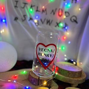 Könnte beinhalten: Ein klares, becherförmiges Trinkglas mit einem roten herzförmigen Aufdruck mit der Aufschrift "VECNA/ HENRY/ 001". Das Glas steht auf einem Holzuntersetzer, mit bunten Lichterketten und einem weißen Hintergrund mit Buchstaben.