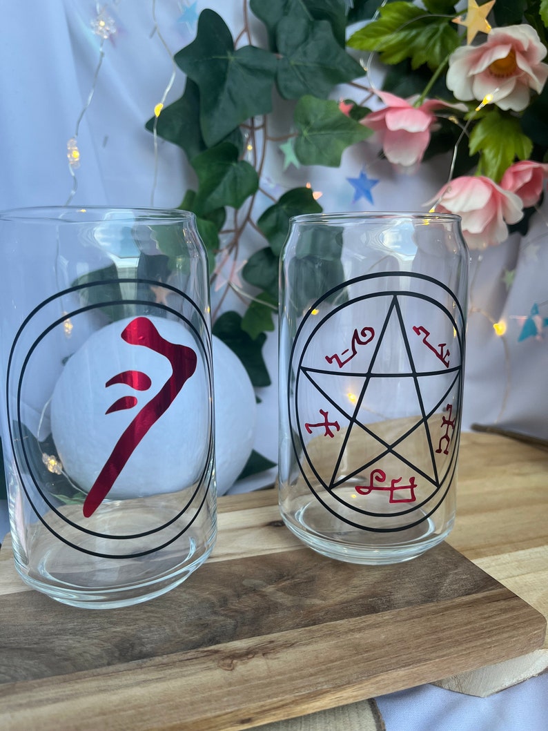 Pu&ograve; includere: Due bicchieri trasparenti a forma di lattina con grafiche nere e rosse. Uno presenta un simbolo rosso all'interno di un cerchio nero, l'altro un pentagramma con simboli rossi. Appoggiati su una superficie di legno.
