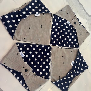 May include: Four square fabric coasters. Each coaster features a navy blue polka dot pattern on one side and a tan linen-like fabric on the other. The tan side has small black triangle designs. Each coaster has a snap closure.