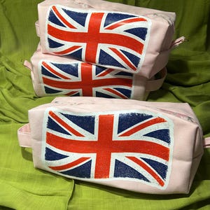 May include: Three pink wash bags, each featuring a Union Jack flag design. The flag has a red cross and diagonal stripes on a white background, set against a blue background. Each bag has a zip closure and a small handle.