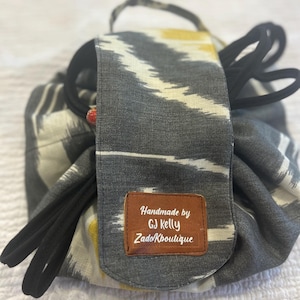 May include: A grey and white patterned cosmetic bag with a black drawstring closure. The bag has a handle and a flap with a brown leatherette tag that reads "Handmade by GJ Kelly ZadoKbouitique."