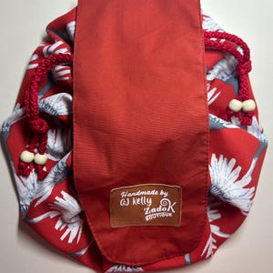 May include: Red drawstring bag with a floral pattern in white and grey. The bag has a solid red flap and red drawstring with white beads. The bag has a brown leather tag that says "Handmade by GJ Kelly ZadoK Boutique."