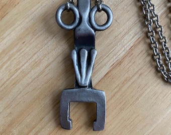 May include: A silver pendant necklace with a unique abstract design. The pendant features a stylized figure with a rounded head and two circles for eyes. The pendant is attached to a silver chain.
