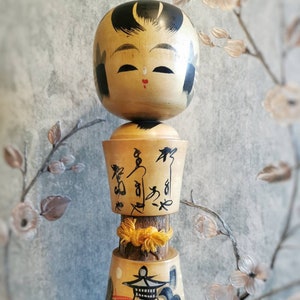 Può includere: Una bambola di legno giapponese tradizionale, conosciuta come Kokeshi, con il viso dipinto e i capelli neri. La bambola è impilata su altri due cilindri di legno, uno con caratteri giapponesi e l'altro con una scena dipinta di un pagoda e onde.