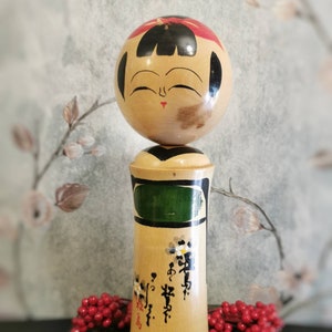 Può includere: Una bambola di legno giapponese tradizionale con la testa rotonda e il viso dipinto. La bambola indossa un kimono verde e bianco con motivi floreali in bianco e nero e caratteri giapponesi.