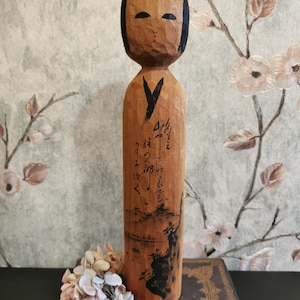 Può includere: Bambola Kokeshi in legno con viso intagliato, capelli neri e un accento rosso. Il corpo cilindrico presenta calligrafia nera e un dipinto di paesaggio. La bambola è alta circa 25 cm, mostrando l'artigianato tradizionale giapponese.