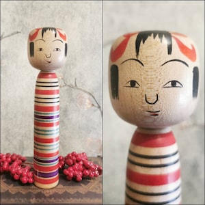Può includere: Una bambola di legno giapponese tradizionale con un vestito a righe rosso, bianco e blu. La bambola ha un viso dipinto con capelli neri e accenti rossi.