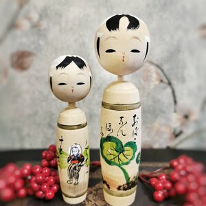 Può includere: Due bambole di legno giapponesi tradizionali, conosciute come kokeshi, con volti dipinti e motivi floreali. La bambola più grande ha il testo "お は よ う" scritto sul suo corpo.